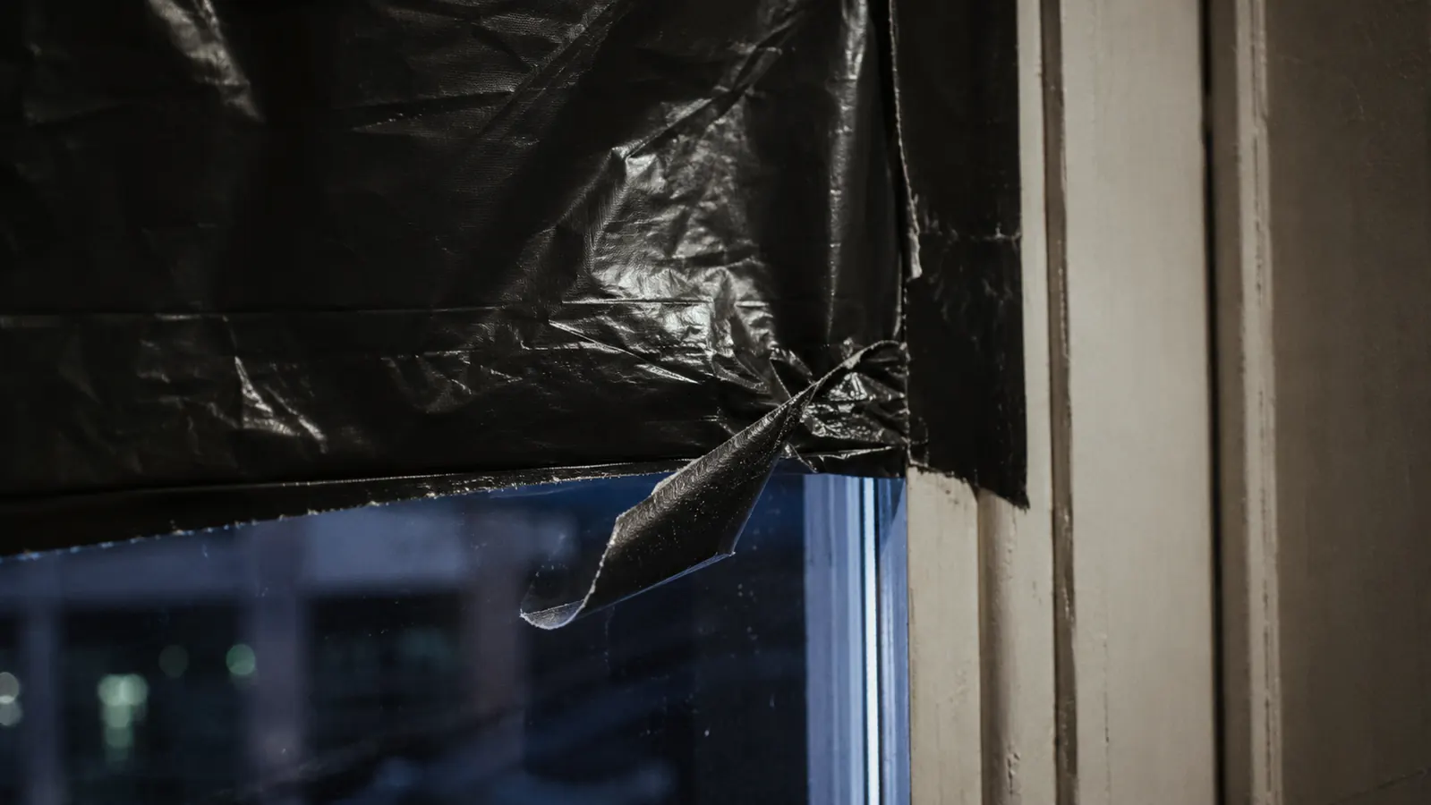 A corner of dark plastic bin liner taped to a window pane with the tape starting to peel