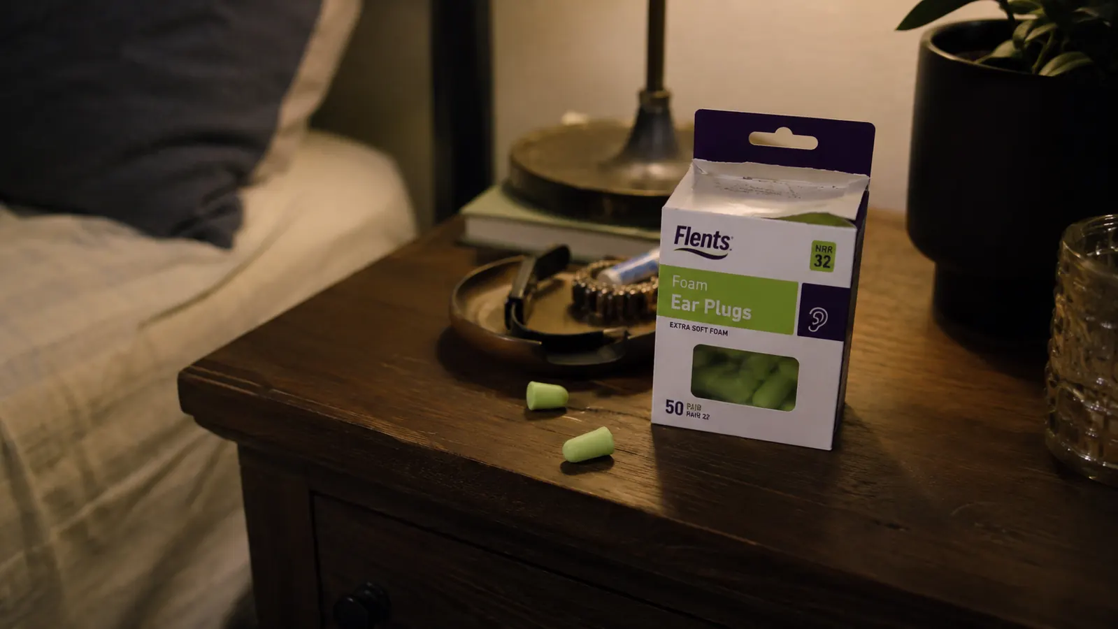 Small green box of Flents foam ear plugs sitting open on a nightstand next to a lamp and a glass of water