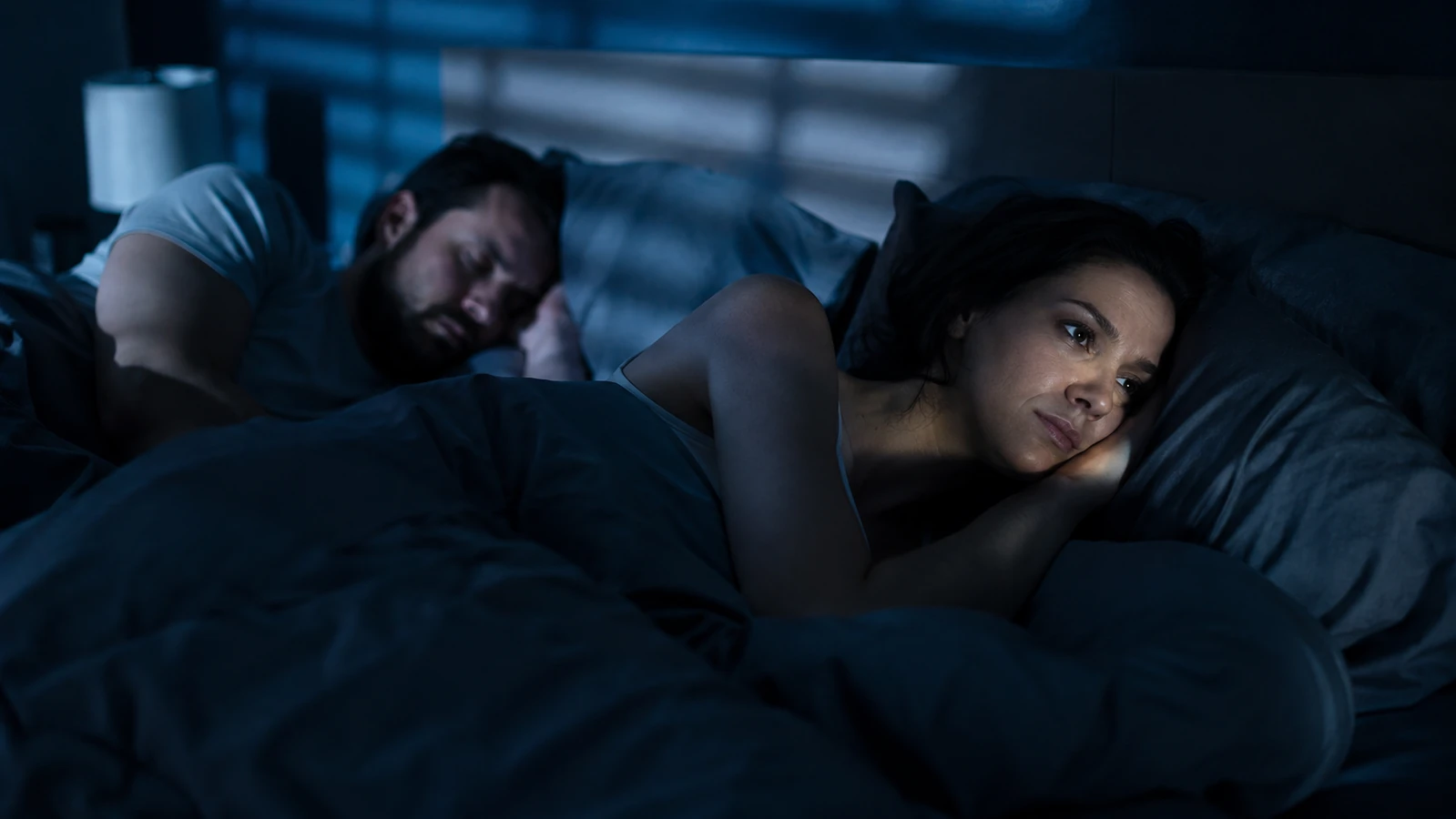 Woman lying awake in bed at night, eyes open, staring at the ceiling next to her sleeping partner