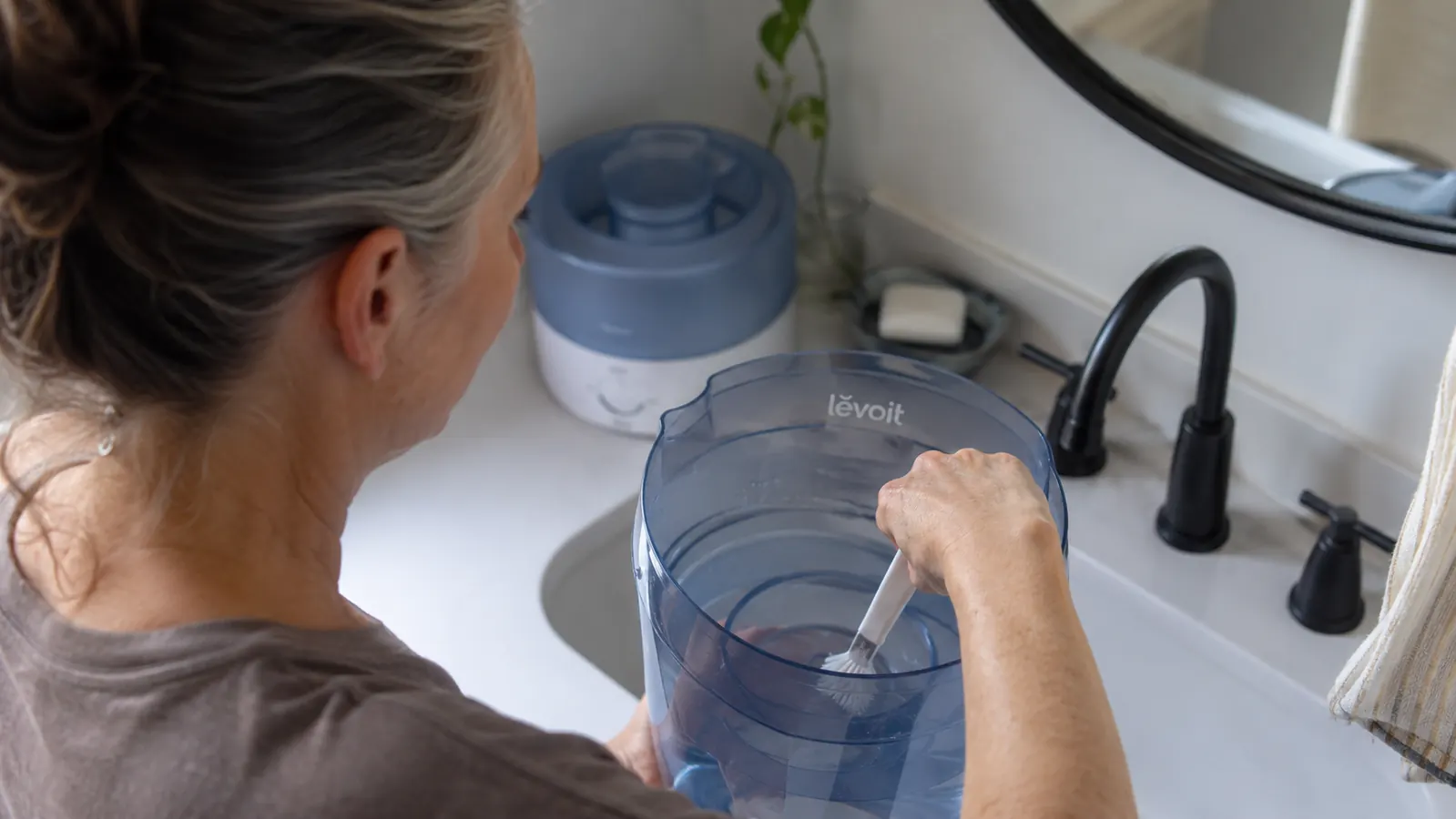 Hand using a small brush to scrub mineral scale from inside a humidifier tank over a bathroom sink