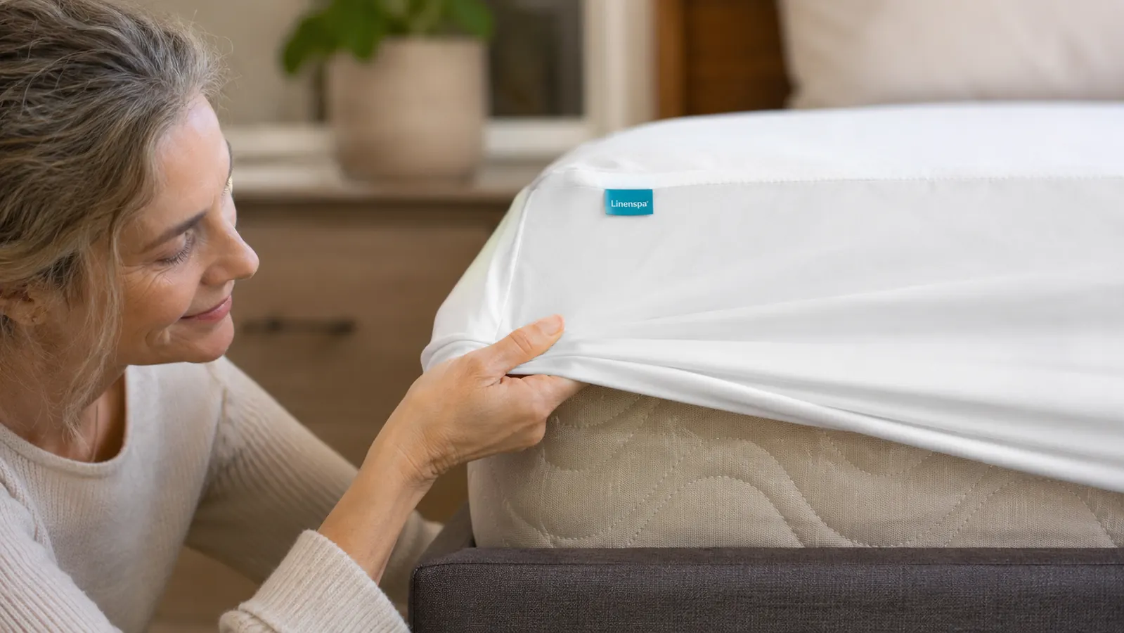 Hand pulling the elasticized corner of a white waterproof mattress protector over a bare mattress corner