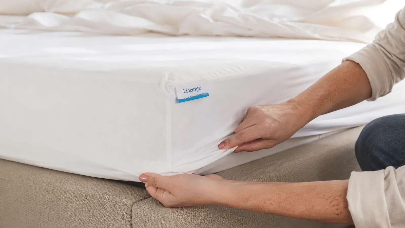 Hands stretching the fitted elastic corner of a white waterproof mattress protector over a bare mattress corner